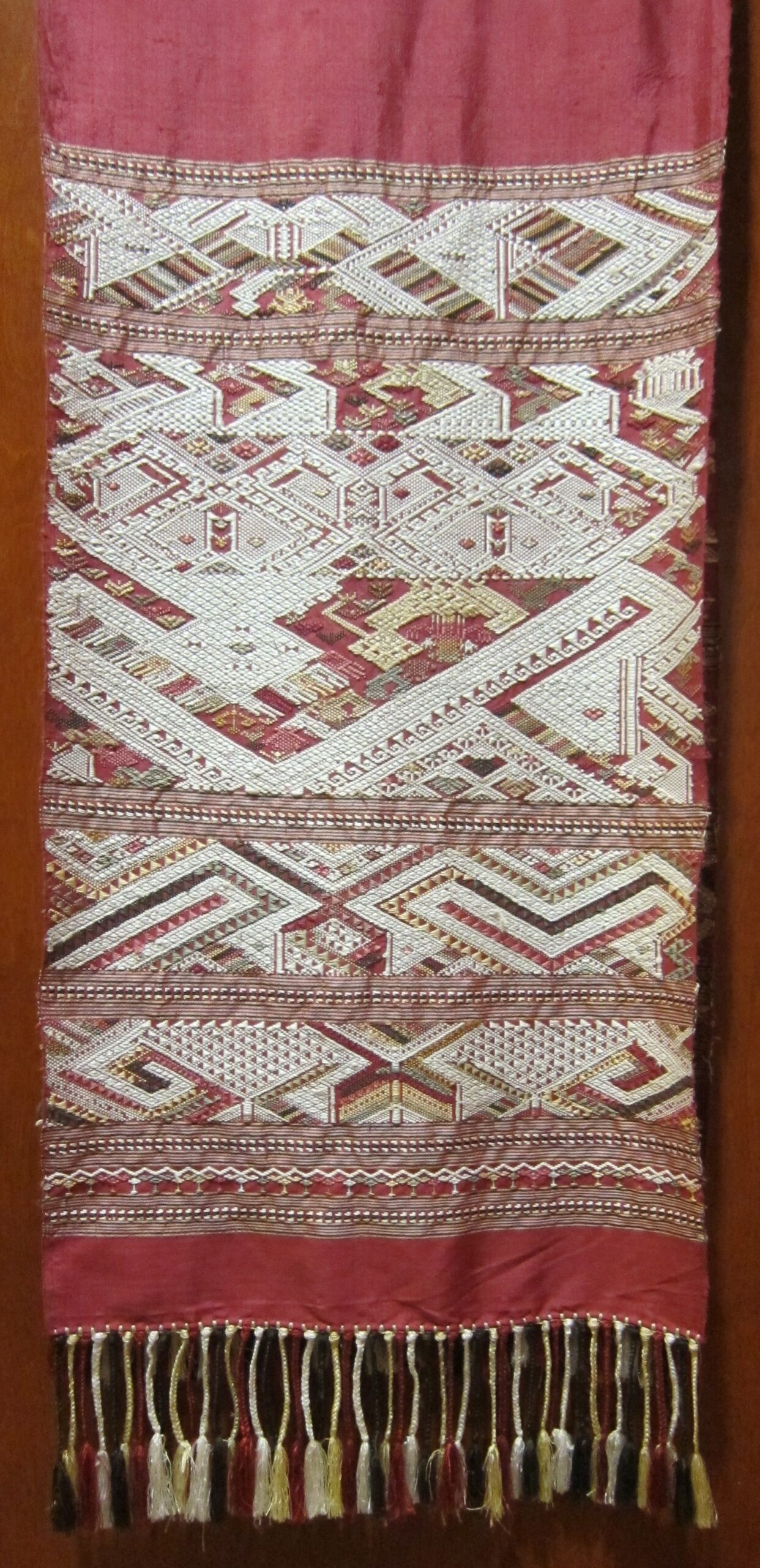 Red Lao silk textile with dense cream and gold geometric woven bands and a tasseled lower edge